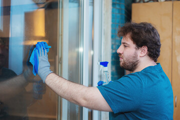 A Caucasian bearded young man washes the window in the kitchen.