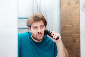 A bearded Caucasian young man shaves in the bathroom.