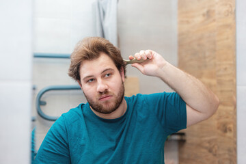 A bearded young Caucasian man combs his hair in the bathroom.