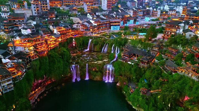 Aerial view of Furong Ancient Town's architecture alongside illuminated waterfalls cascading into a dark pool, Xiangxi Tujia, China.