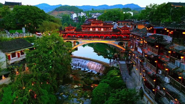 Aerial view of a vibrant bridge adorned with red lanterns, reflecting on the water, surrounded by lush greenery and traditional buildings, Xiangxi, China.