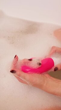 Female hand lowers pink sex toy onto soap suds filling tub, evoking private bathing ritual with added stimulation for ultimate relaxation and personal indulgence