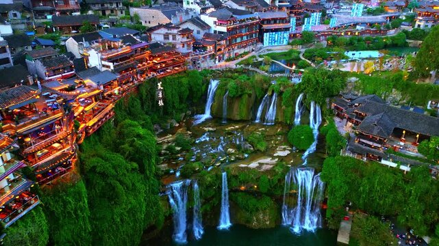 Aerial view of waterfalls cascading into a pool, juxtaposed with traditional houses illuminated with warm lights, creating a stunning contrast, Xiangxi Tujia, China.