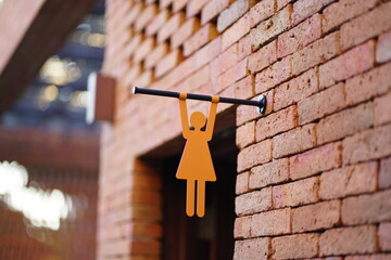 Women&rsquo;s restroom symbol designed as a woman hanging from a horizontal bar on a brick wall, blending wayfinding signage with themes of female strength, patience, resilience, and urban life.