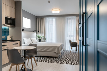 Fototapeta premium modern studio apartment design. apartment has large double bed, desk and chair, large windows with curtains, built-in wooden kitchen with blue tiles and dark countertop. houseplant next to bed.