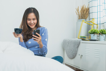 Happy young Asian woman sitting on bed, holding a coffee cup and smiling while looking at her smartphone in a bright cozy bedroom, morning lifestyle, social media, and digital connectivity 