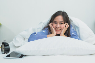 Beautiful happy young Asian woman lying on white bed with chin on hands in cozy bright bedroom, smiling at camera with tablet and alarm clock nearby, morning lifestyle and wellness concept at home.