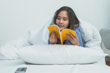 Happy young Asian woman lying on white bed and reading an interesting yellow book in a bright cozy bedroom, morning home lifestyle, education and hobby concept.