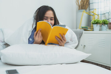 Happy young Asian woman lying on white bed and reading an interesting yellow book in a bright cozy bedroom, morning home lifestyle, education and hobby concept.