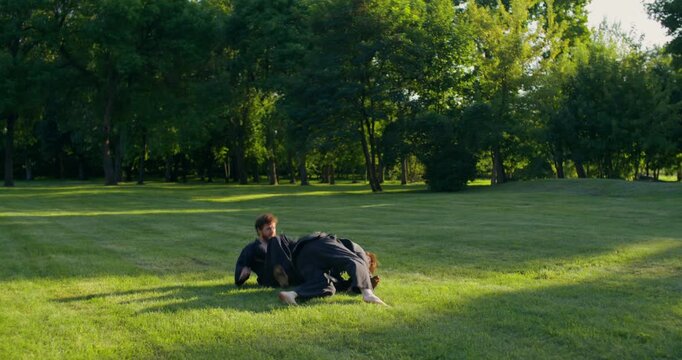 Male karate practitioners fight and fall onto the grass during a training match. The karate practitioners put on black keikogi and prepare to train.