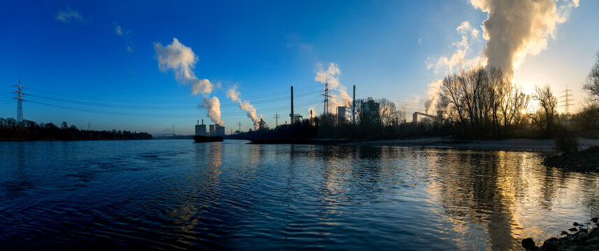 Panorama of a large steelworks in Duisburg-H&uuml;ttenheim Ehingen reflected in calm water surface of Rhine River. Heavy emissions and steam in the clear blue sky on a cold winter morning at sunrise.