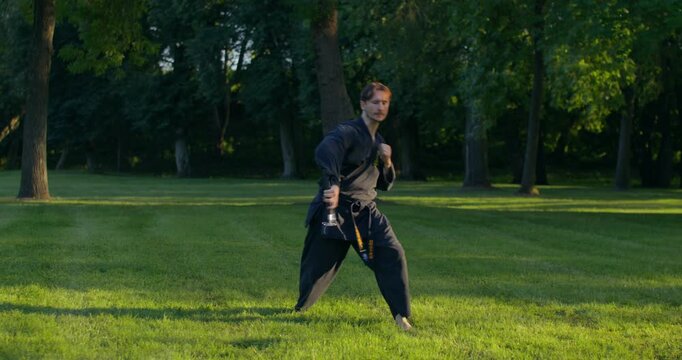 A male karateka assumes a starting position and then begins training with nunchaku. A male karateka trains on the grass with nunchaku and tonfa.
