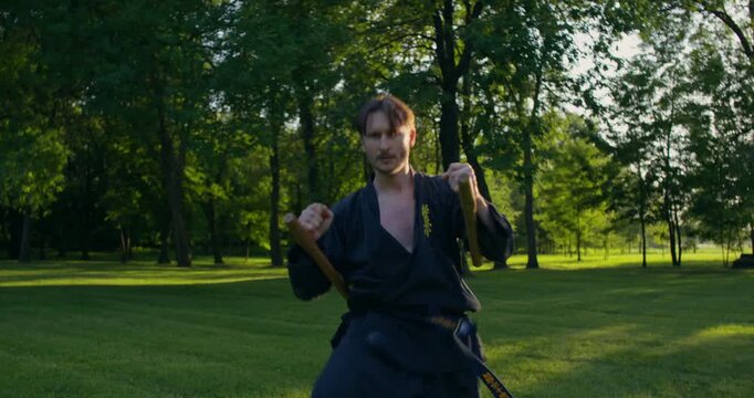 Male karateka training to combine tonfu and leg strikes. A male karateka trains on the grass with nunchaku and tonfa.