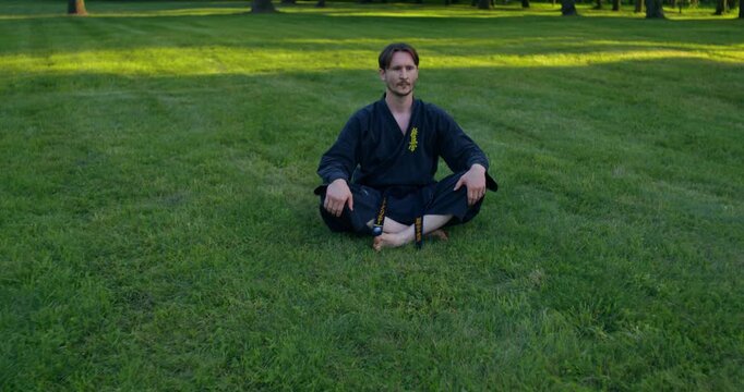 Male karateka sitting cross-legged on the grass. A male karateka trains on the grass with nunchaku and tonfa.