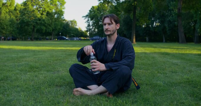 Male karateka opening a bottle and drinking water while sitting on the grass after training. A male karateka trains on the grass with nunchaku and tonfa.