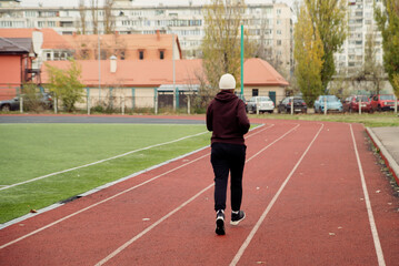 Fototapeta premium Rear view of a person in athletic wear walking on a red running track at an outdoor stadium during autumn. Focus on health, exercise, and urban fitness activity.