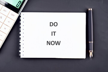 A notebook displays DO IT NOW in bold lettering. Placed on a dark surface with a calculator and a pen, urging immediate action and emphasizing the present