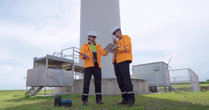 Two engineers in orange uniform at wind turbine tower analyze operation report verify technical data coordinate maintenance ensuring safe renewable electricity performance and clean energy production