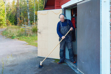 Everyday maintenance as an older man sweeping the ground outside a workshop, steady movement and simple task expressing continued activity after 60.
