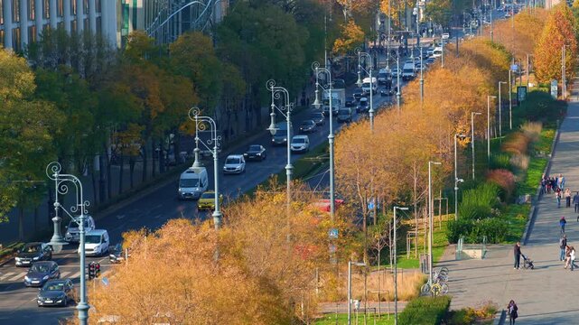 view of a busy Budapest street in autumn, featuring historic architecture, yellow fall trees, city traffic with cars and buses, and pedestrians walking on sunny sidewalks