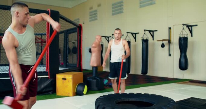 Two male mixed martial artists work on their striking power using a sledgehammer and a tire.. One man trains another in the ring.
