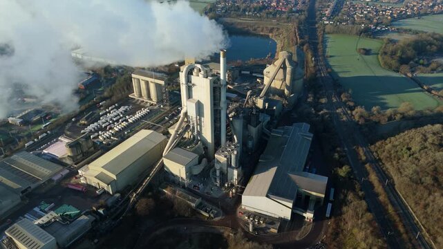 Aerial view of the large cement plant billowing smoke near Lawford Road, juxtaposed against a lake and green fields, Rugby, England, United Kingdom.