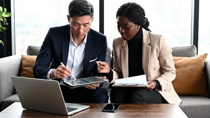 Diverse business professionals collaborate, discussing strategy using a tablet and laptop in a modern office lounge. Focused colleagues analyze data, planning for financial success.