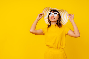 Beautiful happy young Asian woman in vibrant yellow outfit wearing a wide-brimmed straw hat and white sunglasses, smiling cheerfully against a solid yellow background, summer vacation 