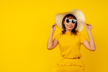 Beautiful happy young Asian woman in vibrant yellow outfit wearing a wide-brimmed straw hat and white sunglasses, smiling cheerfully against a solid yellow background, summer vacation 