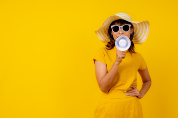 Beautiful happy young Asian woman in vibrant yellow outfit with straw hat and sunglasses holding a megaphone, shouting and making an announcement on isolated yellow background