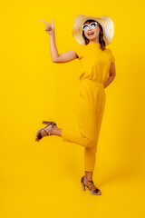Full body portrait of a happy young Asian woman in a vibrant yellow jumpsuit and straw hat, pointing up at copy space with a cheerful smile, summer fashion and travel vacation concept 