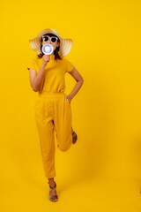Beautiful happy young Asian woman in vibrant yellow outfit with straw hat and sunglasses holding a megaphone, shouting and making an announcement on isolated yellow background