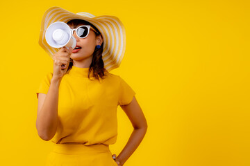 Beautiful happy young Asian woman in vibrant yellow outfit with straw hat and sunglasses holding a megaphone, shouting and making an announcement on isolated yellow background