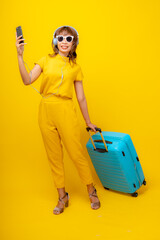 Cheerful young Asian woman in vibrant yellow jumpsuit and straw hat crouching on isolated yellow background, smiling at camera, summer vacation and fashion travel concept.