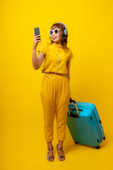 Cheerful young Asian woman in vibrant yellow jumpsuit and straw hat crouching on isolated yellow background, smiling at camera, summer vacation and fashion travel concept.