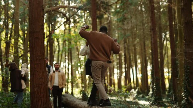 A group of people are walking on a log in a forest. One man is holding a bag