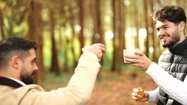 Two men are standing in a forest, one of them is holding a cup. They are smiling and seem to be enjoying each other's company