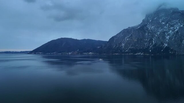 Aerial drone rising forward from land over alpine lake with boat and snowy mountain winter landscape Traunsee Austria