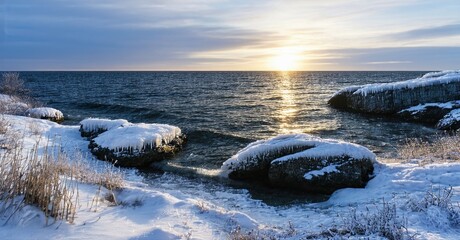 Winter scene. Rocks covered snow near ocean. Sun shining through clouds creates golden reflection water. Calm, serene landscape.