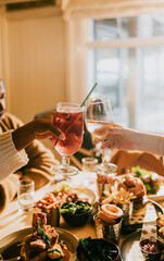 Naklejka premium A group of people are celebrating with drinks and food. They are holding up their glasses and toasting to each other. The table is filled with various dishes, including a plate of fries and a sandwich