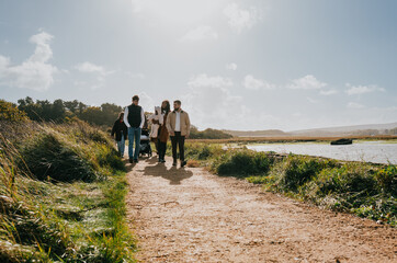Obraz premium A family of four is walking along a dirt path near a body of water. The path is lined with grass and there are trees in the background. The family appears to be enjoying a leisurely walk together