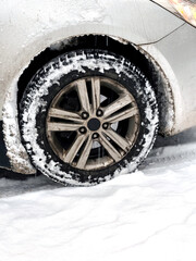Close up of a dirty car wheel with alloy rim and winter tire covered in snow. Vehicle parked on snowy ground, visible tread pattern, ice, slush, and snow buildup around the wheel arch.