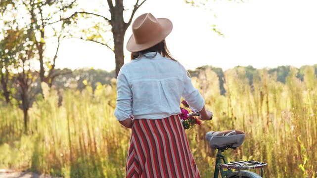 A woman walks beside her bicycle on a path lined with trees and tall grass in the afternoon light. She wears a hat and a striped skirt