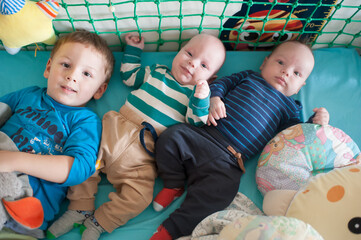 Three boys, including two babies, are lying side-by-side on a turquoise surface. They are surrounded by colorful stuffed animals. The area is enclosed by green netting
