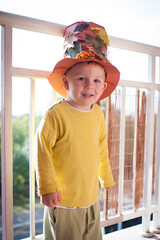 A cute young boy stands on a balcony wearing a homemade hat decorated with leaves and flowers. He is smiling and seems to be having fun on a bright day