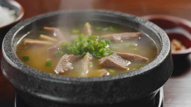 Steaming hot japanese gyutan soup (ox tongue) or savory asian beef stew served in a black stone bowl garnished with fresh green onions.