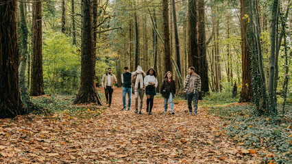 Fototapeta premium A group of five people are walking through a forest, surrounded by trees and leaves. The atmosphere is calm and tranquil, as they enjoy the beauty of nature. The group consists of a mix of ages