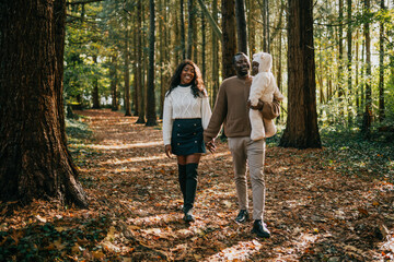 Fototapeta premium A couple holding a baby in a forest. The baby is wearing a white jacket. The couple is walking on a path in the woods