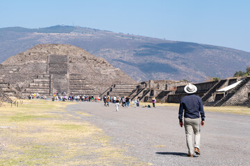 Man in a hat walking toward the Pyramid of the Moon at the Teotihuac&aacute;n archaeological site in Mexico under a clear blue sky.