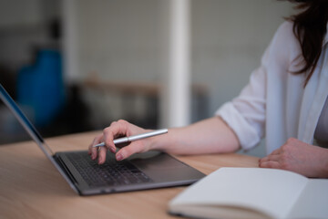 Woman's hand holding a pen while typing on a laptop keyboard, working remotely from a wooden desk...
